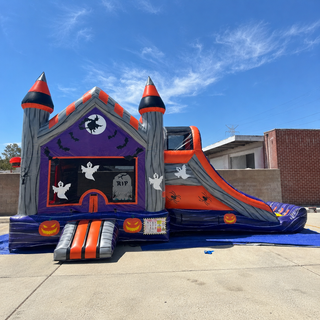 A Halloween-themed inflatable bounce house and slide set up outdoors on a concrete patio under a blue sky. The castle-style bounce house features gray walls, orange roofs, purple accents, white ghost cutouts, a moon and witch graphic, bats, tombstone decorations, and a side slide with orange and gray panels.