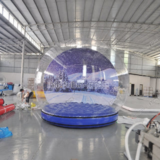 A large inflatable transparent bubble with a printed snowy mountain landscape and a blue base, photographed inside a warehouse. A person is visible working at the left edge, and the warehouse floor and metal roof are visible in the background.