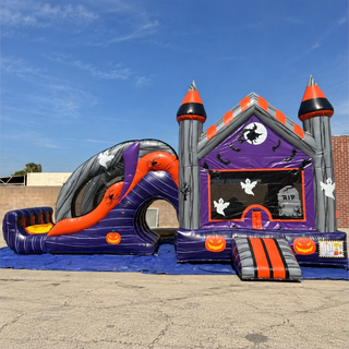 A Halloween-themed inflatable bounce house and slide set set up outdoors on a concrete ground under a blue sky. The bounce house features a purple and gray castle design with orange accents, white ghosts, and tombstone graphics, alongside a matching slide.