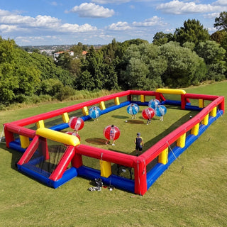An inflatable outdoor foosball court with red, blue, and yellow walls, set up on a grassy lawn with trees and a blue sky in the background. Several people are inside colorful transparent balls playing the game.
