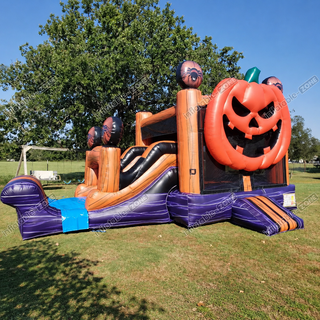 A Halloween-themed inflatable bounce house and slide set on a grassy field, featuring a large orange jack-o'-lantern face, pumpkin decorations, and purple and brown wooden-style pillars. Trees and blue sky are visible in the background.